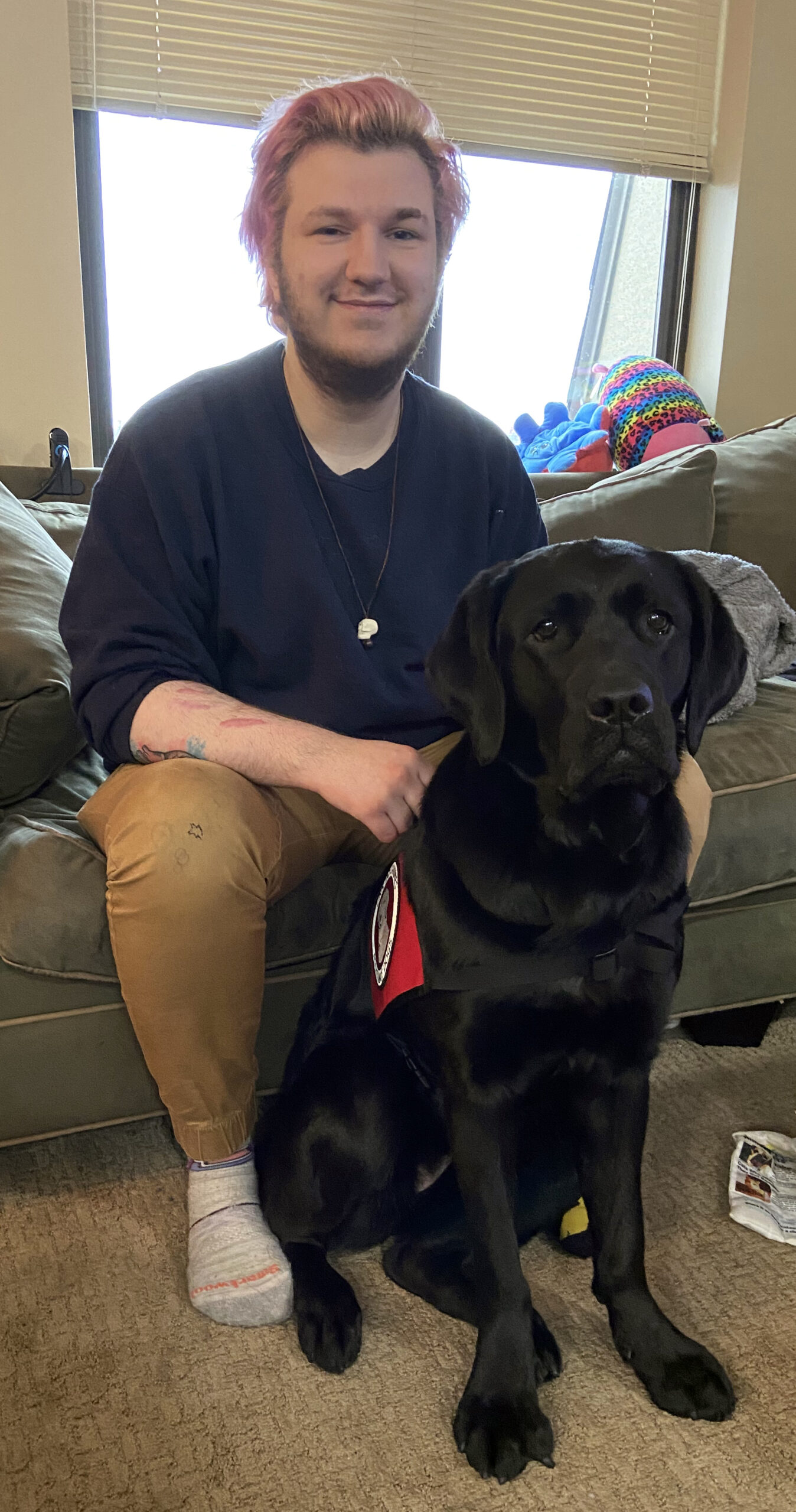 young man sitting on couch with black Lab service dog sitting in front of him
