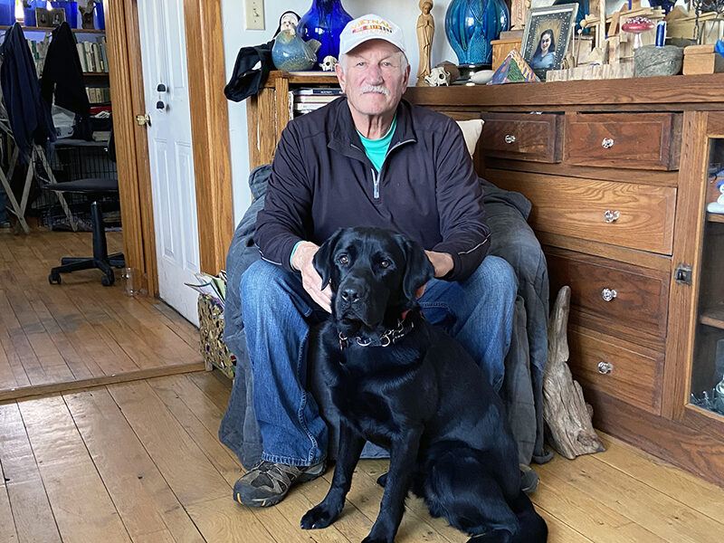 man sitting on chair in living room with black Lab sitting on front between his legs