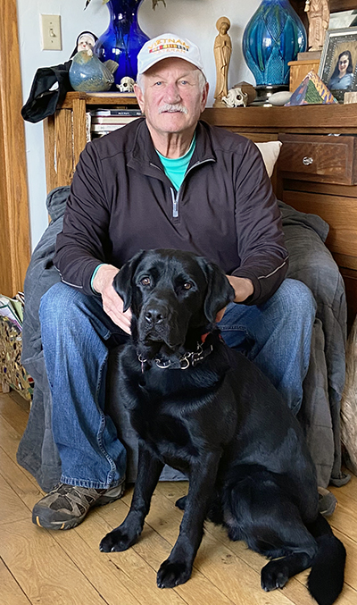 man sitting on chair in living room with black Lab sitting on front between his legs