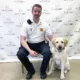male paramedic sitting on chair next to sitting yellow Lab in front of Can Do Canines backdrop