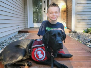 boy sitting on front sidewalk with black Lab dog wearing Can Do Canines assistance dog cape