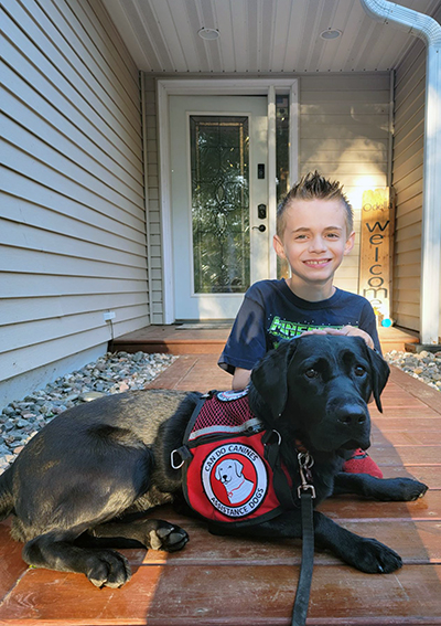 boy sitting on front sidewalk with black Lab dog wearing Can Do Canines assistance dog cape