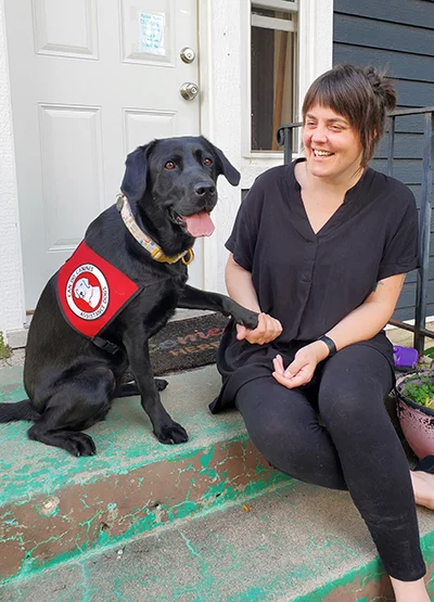 woman sitting on front step looking at black Lab dog wearing Can Do Canines service cap sitting next to her