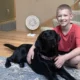 Boy in red shirt sitting on floor inside home with arm around black Lab dog