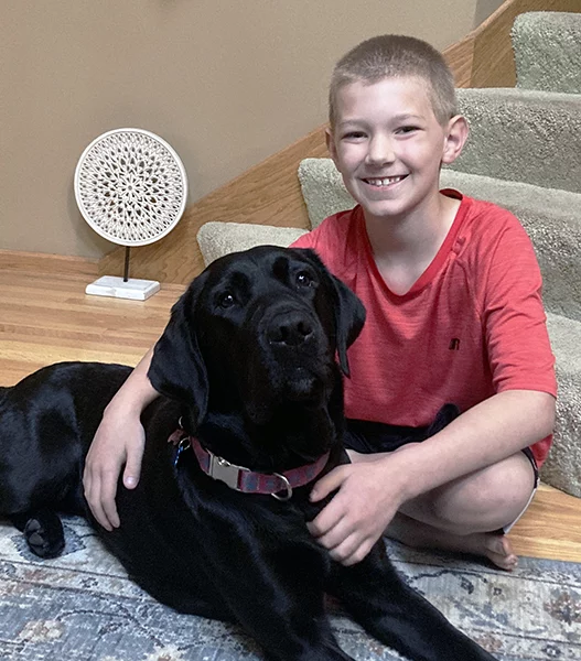 Boy in red shirt sitting on floor inside home with arm around black Lab dog