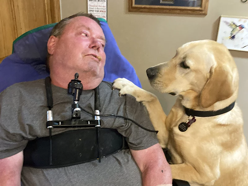 man sitting in wheelchair with yellow Lab dog sitting next to him and putting paw on his shoulder