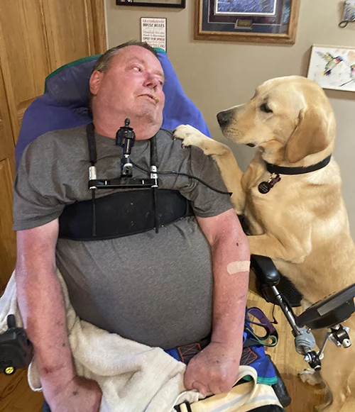 man sitting in wheelchair with yellow Lab dog sitting next to him and putting paw on his shoulder