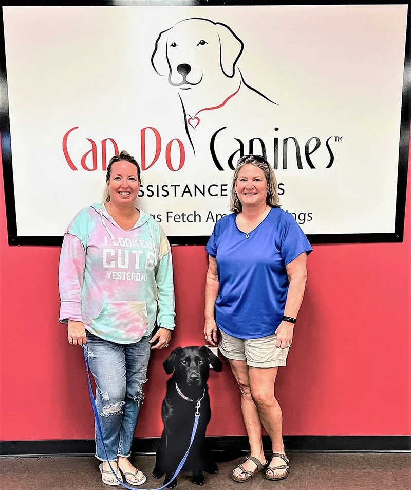 Two women and black Lab standing in front of Can Do Canines sign