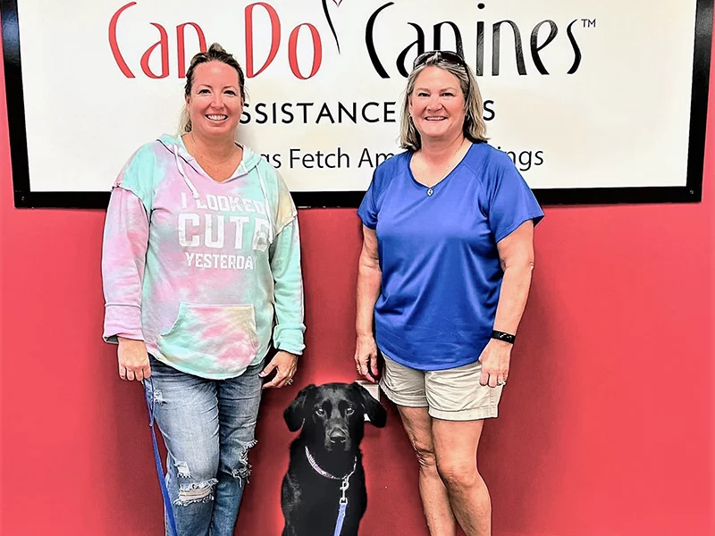 Two women and black Lab standing in front of Can Do Canines sign