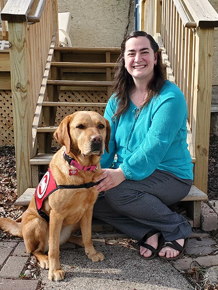 woman kneeling at bottom of home stairs next to yellow Lab dog wearing Can Do Canines service cape