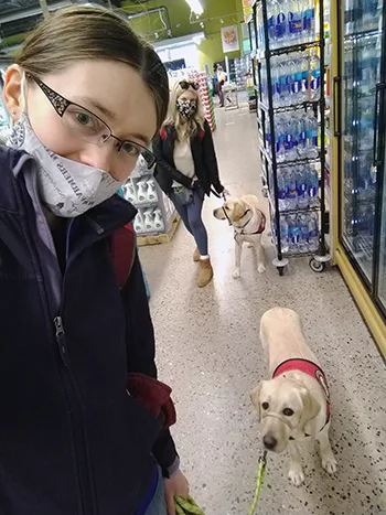 young woman wearing facemask in grocery store with yellow Lab service dog and another woman and service dog in background