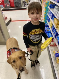 Young boy holding an Oreo pack is stading with his yellow Lab service dog in a grocery aisle.