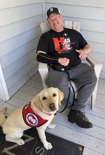 man sitting in rocker on porch with yellow Lab sitting in front of him wearing Can Do Canines service cape