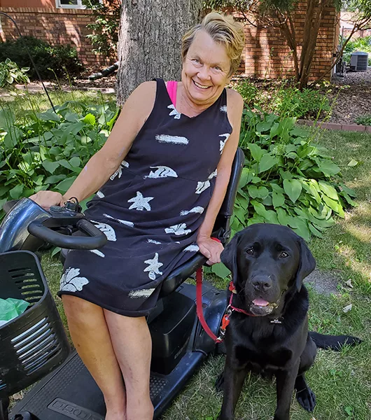Woman sitting in powerchair, with black Lab sitting on lawn beside her