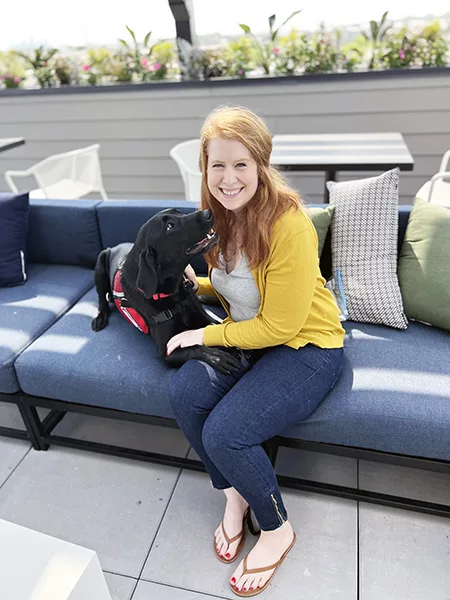 young woman sitting on outdoor sofa with black Lab service dog lying next to her