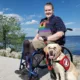 young man sitting in wheelchair near large lake in summertime with yellow Lab service dog sitting on pavement next to him wearing Can Do Canines service cape