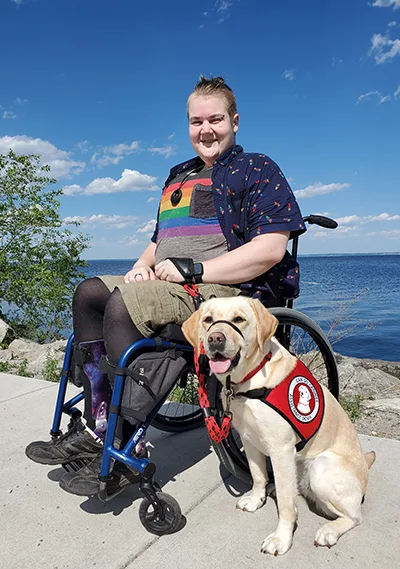 young man sitting in wheelchair near large lake in summertime with yellow Lab service dog sitting on pavement next to him wearing Can Do Canines service cape
