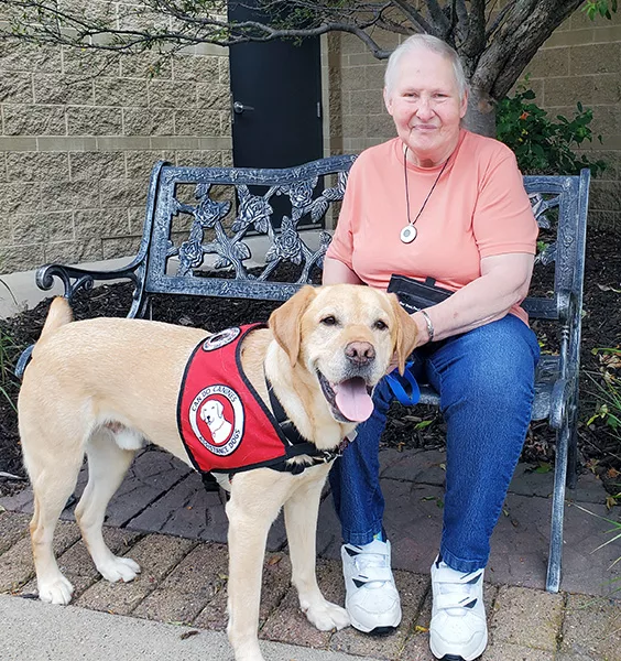 woman sitting on outside bench with yellow Lab service dog wearing Can Do Canines vest standing in front of her