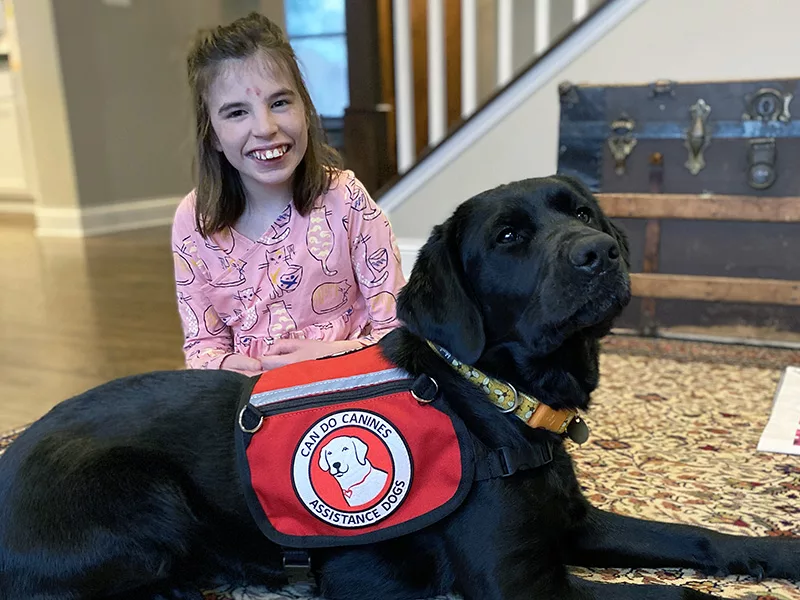 Girl sitting on area rug with black Lab service dog lying in front of her wearing Can Do Canines cape
