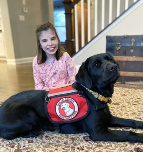Girl sitting on area rug with black Lab service dog lying in front of her wearing Can Do Canines cape