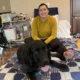 woman sitting on a bed with black Lab dog lying by her legs, with both of them smiling at the camera
