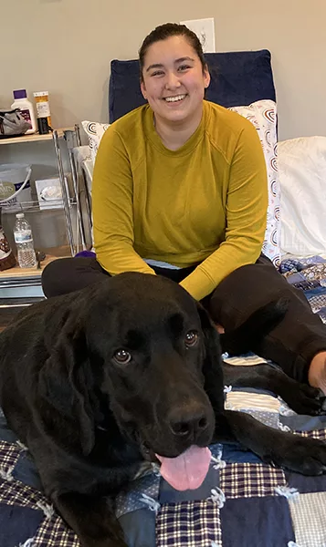 woman sitting on a bed with black Lab dog lying by her legs, with both of them smiling at the camera