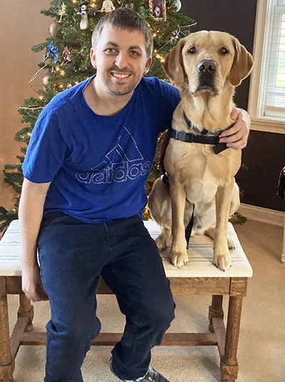 young man and yellow Lab dog both sitting on bench in front of Christmas tree, with dog wearing service cape and man putting arm around dog