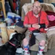 man wearing magnifying glasses and leg braces sitting in plush chair in living room with black Lab dog at his feet looking at him