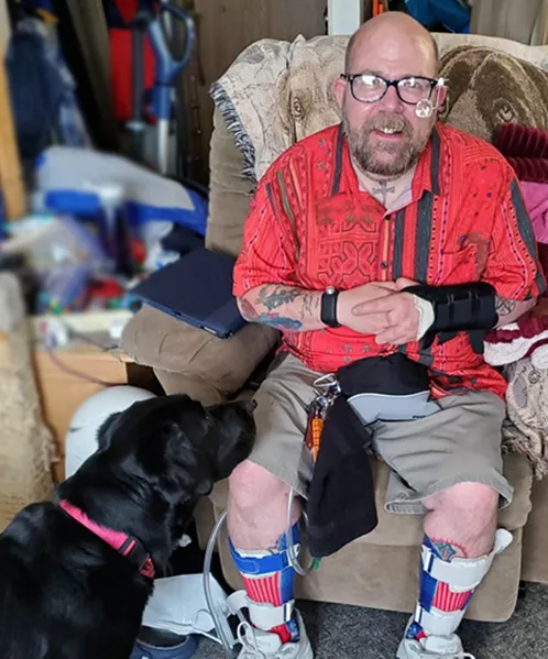 man wearing magnifying glasses and leg braces sitting in plush chair in living room with black Lab dog at his feet looking at him