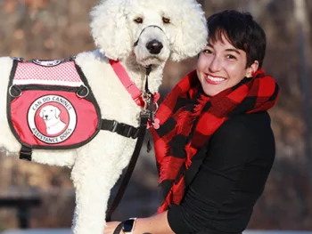 Woman posing with a white Standard Poodle wearing a red Can Do Canines cape