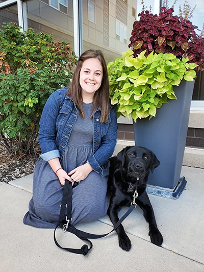 woman kneeling on sidewalk with black Lab dog lying next to her and both are looking at the camera
