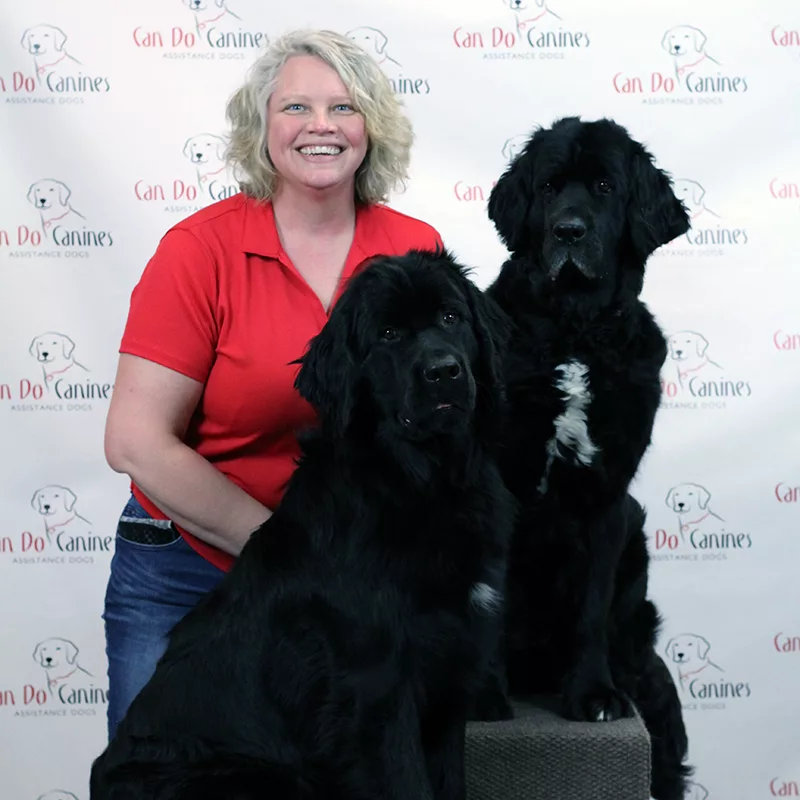 Woman wearing red shirt and smiling at the camera with two very large black dogs sitting with her