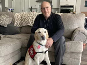 man sitting on sofa, leaning forward to have hands on yellow Lab service dog sitting between the man's feet