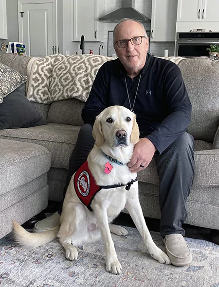 man sitting on sofa, leaning forward to have hands on yellow Lab service dog sitting between the man's feet