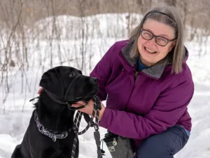 woman kneeling down in snow next to black Lab dog that she has on a leash
