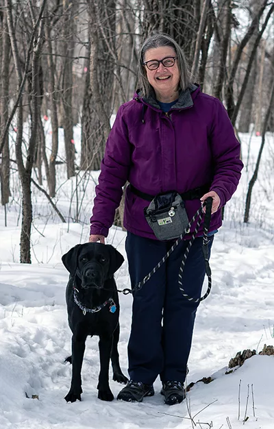 woman standing outdoors in snowy climate with hand on leashed black Lab dog