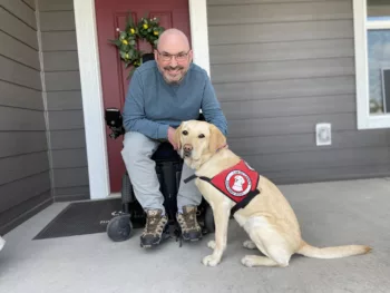 man using a wheelchair is posting with a yellow Lab wearing a red Can Do Canines cape