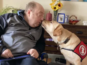 man sitting in wheelchair, leaning to one side to share kiss with yellow Lab service dog wearing Can Do Canines cape