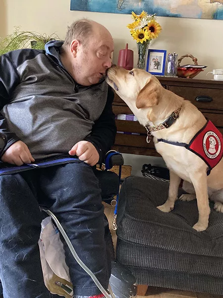 man sitting in wheelchair, leaning to one side to share kiss with yellow Lab service dog wearing Can Do Canines cape