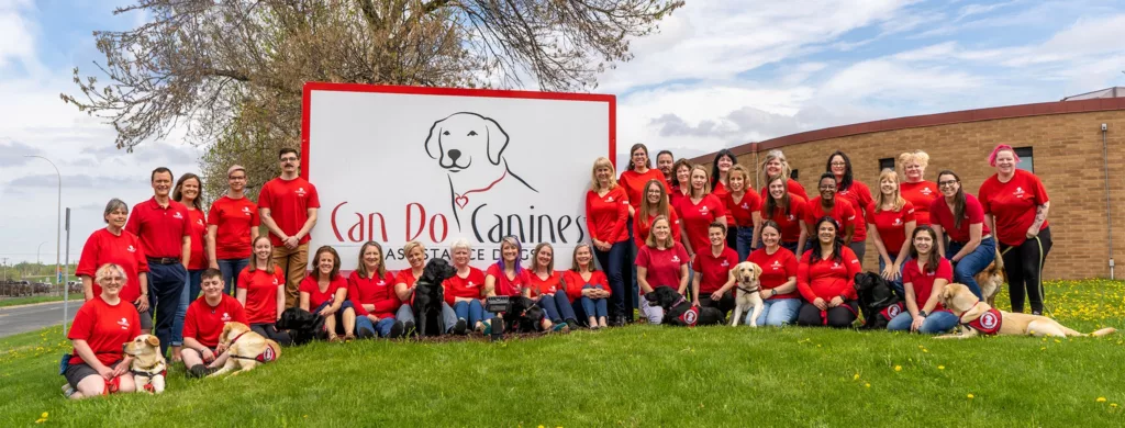 Group of nearly 40 people wearing red Can Do Canines staff shirts are posing in front of the Can Do Canines building.