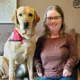 woman with long hair and glasses sitting on sofa with yellow Lab service dog wearing red cape sitting next to her