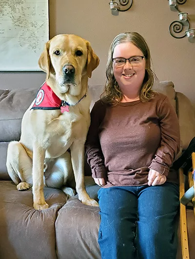 woman with long hair and glasses sitting on sofa with yellow Lab service dog wearing red cape sitting next to her
