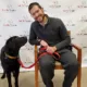 Man, smiling, sitting in chair in front of Can Do Canines backdrop sheet, leaning toward black Lab standing beside him looking at him