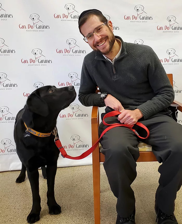 Man, smiling, sitting in chair in front of Can Do Canines backdrop sheet, leaning toward black Lab standing beside him looking at him