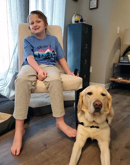 9-year-old blond boy sitting in chair with yellow Lab service dog lying on floor in front of him