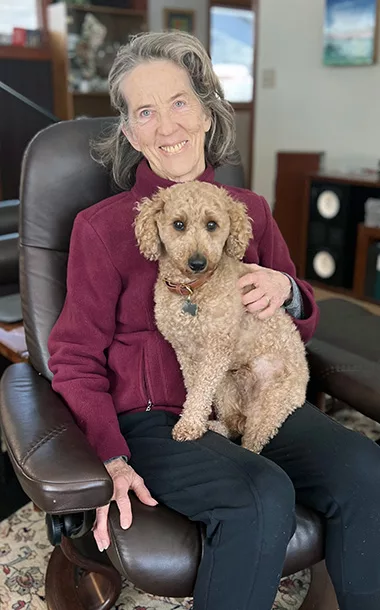 Woman sitting in a chair with small, tan poodle sitting on her lap. Both are looking at the camera.