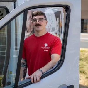 Man wearing a red Can Do Canines staff shirt is posing by a white wan with the Can Do Canines logo on it.