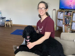 Woman sitting on sofa with black standard poodle with arms around dog and both smiling at camera