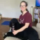 Woman sitting on sofa with black standard poodle with arms around dog and both smiling at camera