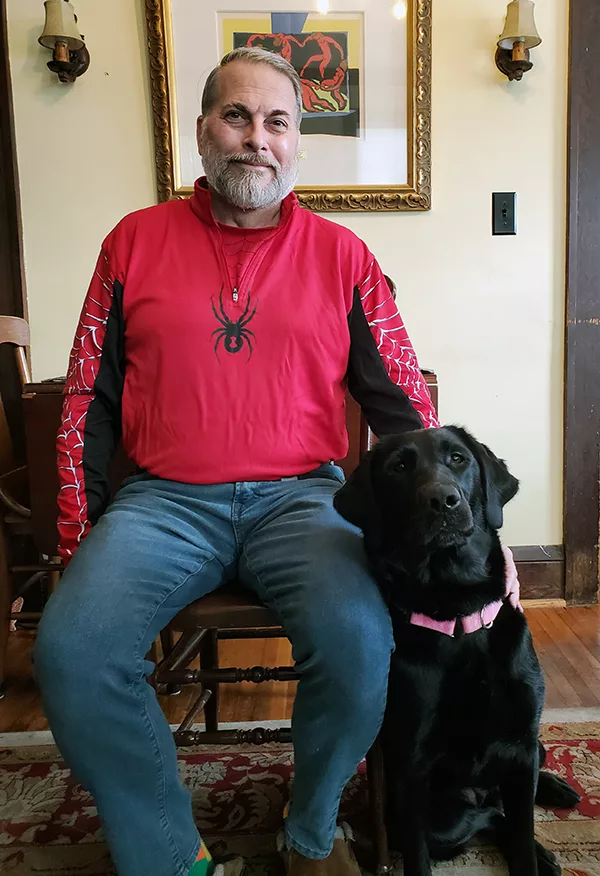 Man sitting on chair with black Lab dog sitting on floor beside him. Both are smiling at camera.